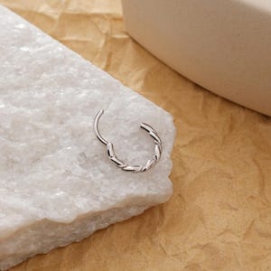 May include: A silver-colored hinged hoop earring with a twisted rope design. The earring is open, revealing the hinge mechanism. The jewelry is set against a white marble surface and a tan background.