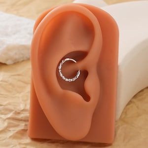 May include: A silver-colored, braided hoop earring is displayed in a simulated ear. The earring is a small, circular design with a textured, woven appearance. The earring is set against a peach-colored ear model.