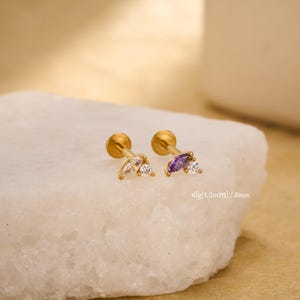 May include: Two gold-coloured, flat-backed earrings with decorative gemstones. One earring features a clear marquise-cut stone and a small round stone. The other has a purple marquise-cut stone and a small round stone. The text "16g (1.2mm) / 8mm" is visible.