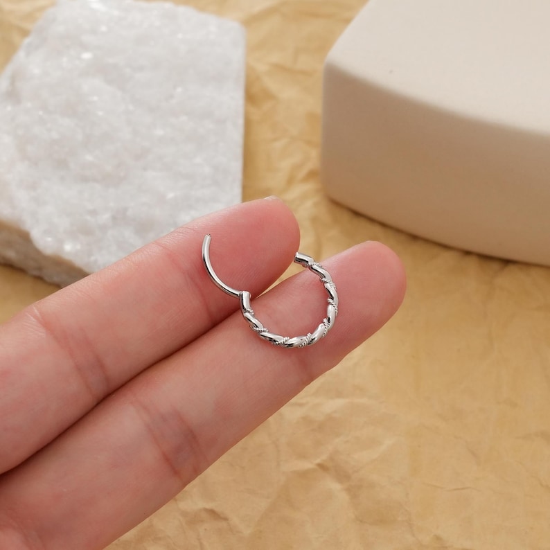 May include: A silver-colored, hinged hoop earring with a twisted rope design. The earring is open, showing the clasp. The jewelry is displayed against a neutral background.