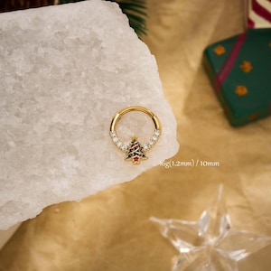 May include: Gold-coloured hinged ring featuring a Christmas tree charm. The charm is red, green, and white. The ring is decorated with clear gemstones. The text on the image reads "16g (1.2mm) / 10mm".