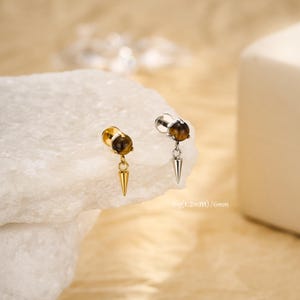 May include: Two ear gauges with a brown stone and a gold or silver spike pendant. The gauges are displayed on a white surface. The text "16g (1.2mm) / 6mm" is visible.