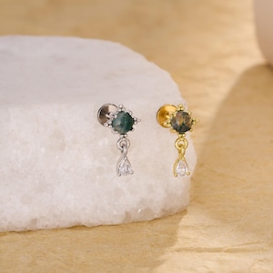 May include: Two flat back stud earrings featuring a round moss agate stone set in a four-prong star-shaped frame with a dangling pear-shaped clear cubic zirconia charm. One earring is silver-toned, and the other is gold-toned metal.