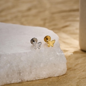 May include: Two butterfly-shaped earrings. One is silver-colored, and the other is gold-colored. The earrings are displayed on a white, textured surface. The text "16g (1.2mm) / 6mm" is visible.