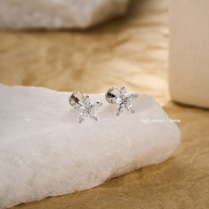 May include: Two silver flower earrings, each with clear, sparkling stones. The earrings are presented on a white surface against a soft, neutral backdrop. The image text reads: 16g (1.2mm) / 8mm.