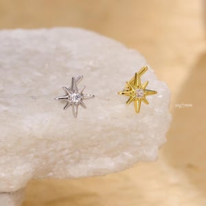 Könnte beinhalten: Zwei sternförmige Ohrringe, einer silberfarben und einer goldfarben, jeweils mit einem zentralen, klaren Edelstein. Der silberne Ohrring befindet sich links, der goldene rechts. Die Ohrringe werden auf einer weißen, strukturierten Oberfläche präsentiert. Text rechts: "20g/7mm".