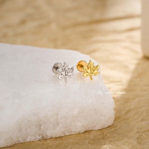 May include: Two maple leaf-shaped earrings, one silver and one gold, are displayed on a white surface. The earrings are small, with a detailed leaf design. The text "16g (1.2mm) / 8mm" is visible.