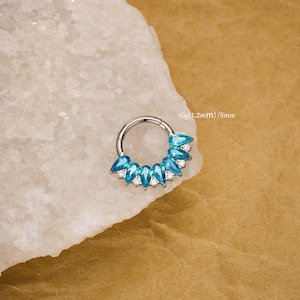 May include: A silver septum ring featuring a series of blue, teardrop-shaped gems and smaller white stones. The ring is 8mm in diameter and 1.2mm thick (16g).
