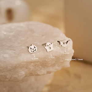 May include: Three silver-colored Halloween-themed stud earrings: a jack-o'-lantern (4.7mm), a ghost (4.2mm), and a bat (3.4mm). The earrings are displayed on a white, textured surface. The gauge is 20g (0.8mm) and the post length is 6mm.