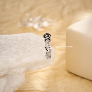 Può includere: Orecchino color argento con un design a rosa. L'orecchino presenta una rosa dettagliata nella parte superiore con accenti neri e uno stelo con foglie. Il testo sull'immagine recita: 14g (1,6 mm)/10 mm.