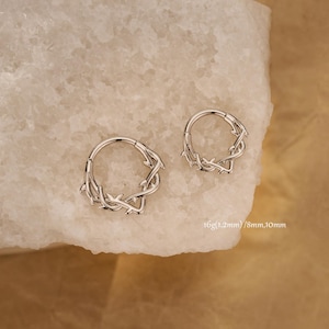 Könnte beinhalten: Zwei silberne Creolen mit einem Dornen-Design. Die Ohrringe werden auf einer hellen Oberfläche gezeigt. Der Text auf dem Bild lautet "16g(1,2 mm) / 8 mm, 10 mm".