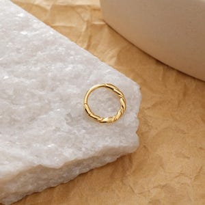 May include: A gold-colored ring with a twisted rope design. The ring is circular and sits on a white, textured surface. The background is a light brown color.