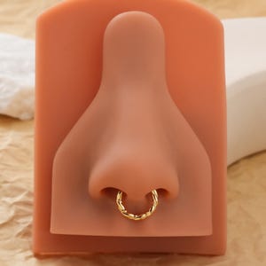 May include: A gold-colored nose ring with a twisted rope design. The ring is displayed on a pink nose-shaped form. The background is a light brown color.
