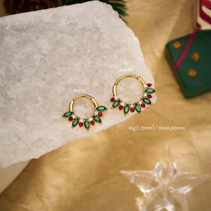 Peut inclure: Deux anneaux de septum dorés, ornés de pierres précieuses vertes et rouges, sont présentés sur une surface blanche. Les anneaux mesurent 8 mm et 10 mm. Le texte "16g(1.2mm) / 8mm,10mm" est également visible.