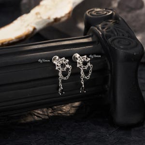 May include: Two silver-toned stud earrings featuring a spiderweb design with a central black stone, a dangling chain, and a teardrop-shaped black stone charm. The left earring is labeled 18g/6mm and the right earring is labeled 18g/8mm.