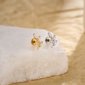 May include: Two leaf-shaped stud earrings. One is gold-toned, the other silver-toned, each adorned with clear, marquise-cut gemstones. The earrings are displayed on a white, textured surface. Text on the image reads: 16g(1.2mm) / 6mm.