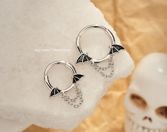 May include: Two silver hoop earrings with black bat wing charms and dangling chains. The earrings are shown on a white surface with a skull in the background. The text on the image reads: 16g(1.2mm) / 8mm,10mm.