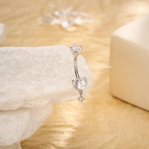 Puede incluir: Un piercing de ombligo plateado con un diseño de barra curva. El anillo presenta un dije en forma de corazón con una piedra preciosa transparente y una piedra preciosa colgante. El texto "14g (1,6 mm)/10 mm" está impreso en el anillo.