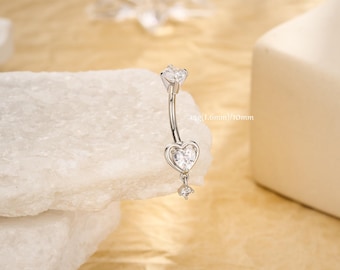 Op de afbeelding: Een zilveren navelpiercing met een gebogen barbell-ontwerp. De ring heeft een hartvormige bedel met een heldere edelsteen en een bungelende edelsteen. De tekst "14g (1,6 mm)/10mm" staat op de ring.