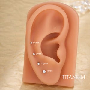 May include: A display of four small, clear, round earrings set in a simulated ear. The earrings are labeled with their sizes: 1.5mm, 2mm, 2.5mm, and 3mm. The word "TITANIUM" is printed in white. The ear is a light peach color.