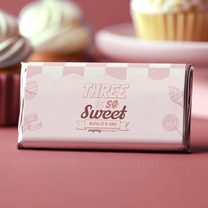 Może przedstawiać: Różowe opakowanie batona z napisem "THREE IS SO Sweet" i "Natalie's 3rd sugary birthday". Opakowanie ma wzór w szachownicę i ilustracje babeczek, makaroników i lizaka.