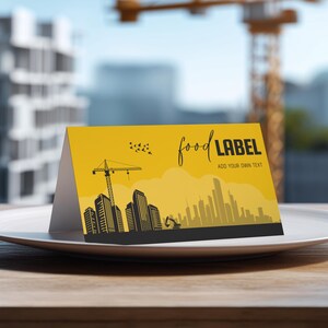 Könnte beinhalten: Eine gelbe Lebensmittelkarten mit einem Stadtbilddesign. Die Karte zeigt eine schwarze Silhouette von Gebäuden, Kränen und den Text "food LABEL ADD YOUR OWN TEXT". Die Karte liegt auf einem weißen Teller.