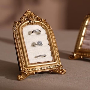Op de afbeelding: Oornate goudkleurige ringdisplay met een crèmekleurig interieur, met drie zilveren ringen met heldere edelstenen. De display heeft een gewelfde bovenkant en decoratieve details.