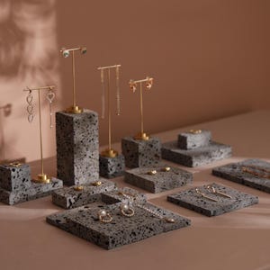 Puede incluir: Una variedad de pendientes dorados, incluyendo modelos de botón y colgantes, presentados sobre bloques de piedra gris texturizada. La exhibición está dispuesta sobre una superficie clara, con iluminación suave que crea sombras.