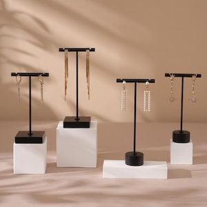 Op de afbeelding: Vier zwarte oorbellen display stands met diverse bungelende oorbellen. De stands staan op witte en zwarte bases. De oorbellen omvatten gouden kettingen, rechthoekige parel ontwerpen en stervormige ontwerpen. De achtergrond is een neutraal beige.