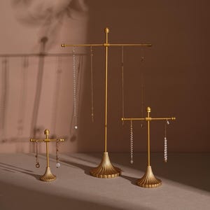 May include: Three gold-colored jewelry display stands of varying heights. Each stand has a decorative base and a horizontal bar for hanging necklaces. The stands are arranged on a neutral surface, with shadows cast on the background.