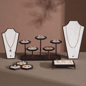 Puede incluir: Una variedad de joyas doradas exhibidas en varios soportes. Las joyas incluyen collares, anillos y pendientes. Los soportes son marrones y blancos, creando un fondo neutro que resalta los detalles de las joyas. La presentación general es elegante y sofisticada.