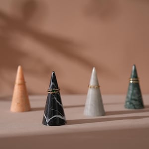 以下が含まれることがあります： ピーチ、黒と白、グレー、緑の色合いの4つの大理石のリングホルダー。各円錐形のホルダーにはリングが表示されています。黒いホルダーには白い縞模様があり、緑のホルダーには金のバンドが付いています。