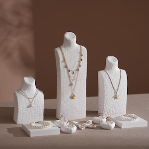 Puede incluir: Exhibición de collares, pulseras y anillos dorados sobre bustos y plataformas blancas. Los collares presentan varios colgantes y diseños en capas. También se ven una pulsera de perlas y anillos, mostrando joyería.
