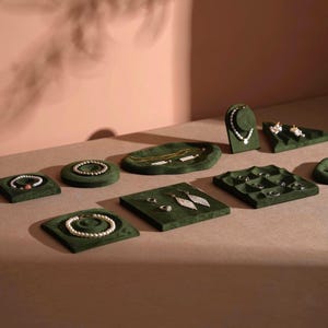Könnte beinhalten: Schmuckpräsentation mit Halsketten, Armbändern, Ohrringen und Ringen. Der Schmuck wird auf grünen Samt-Präsentationsflächen in verschiedenen Formen präsentiert. Der Hintergrund ist eine hellbraune Oberfläche mit einer rosa Wand.