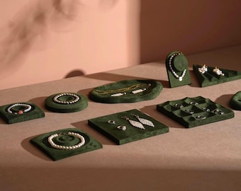 May include: Jewelry display featuring various necklaces, bracelets, earrings, and rings. The jewelry is displayed on green velvet platforms of different shapes. The background is a light brown surface with a pink wall.