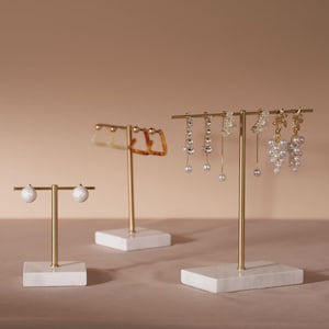 Op de afbeelding: Drie oorbellen display stands met goudkleurige metalen palen en witte marmeren bases. De stands bevatten verschillende oorbellen, waaronder parel studs, bungelende kristallen oorbellen en bloemmotieven. De achtergrond is een effen, neutrale kleur.