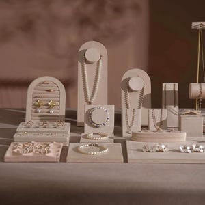 Op de afbeelding: Een collectie beige fluwelen sieradendisplays, waaronder boogvormige kettinghouders, ringplateaus met gleuven, armbandblokken en oorbellenhouders. De set bevat diverse kettingen, parelarmbanden en ringen gerangschikt op de zachte oppervlakken.