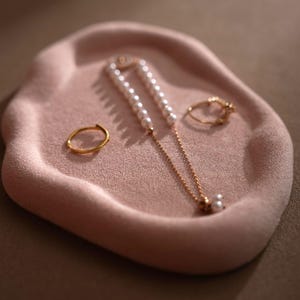 May include: A pink, velvet-like jewelry tray holds a gold ring, a gold necklace with pearls, and a second gold ring. The tray has an organic shape. The jewelry is displayed on a neutral background.