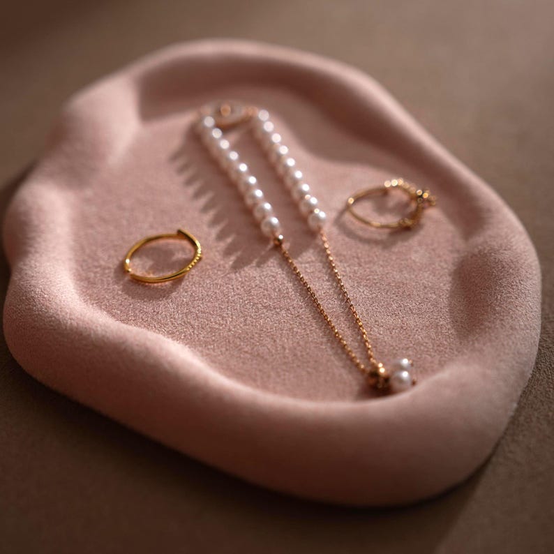 May include: A pink, velvet-like jewelry tray holds a gold ring, a gold necklace with pearls, and a second gold ring. The tray has an organic shape. The jewelry is displayed on a neutral background.