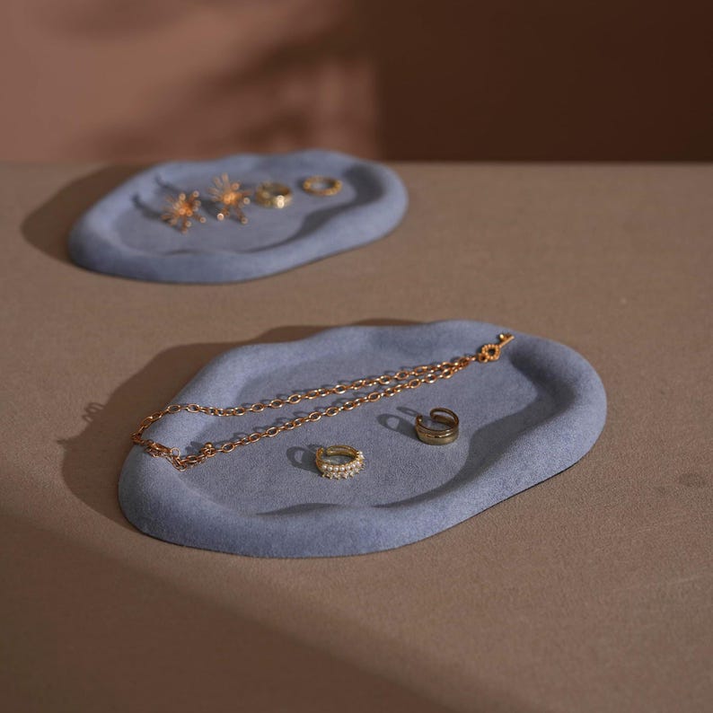 May include: Two light blue, organic-shaped trays with jewelry. One tray holds a gold chain, a pearl-studded ring, and a gold ring. The other tray holds several gold rings and star-shaped earrings. The trays are on a neutral surface.