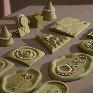 Op de afbeelding: Een assortiment sieraden, waaronder kettingen, armbanden en oorbellen, tentoongesteld op diverse olijfgroene fluwelen displaystandaards. De sieraden zijn voorzien van parels en goudkleurig metaal. De displaystandaards hebben verschillende vormen, waaronder kegels, vierkanten en trays.