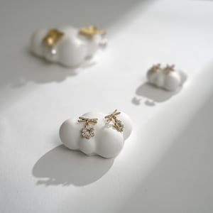 Op de afbeelding: Een paar gouden oorbellen met een klein bloemetjesontwerp en een strikdetail. De oorbellen worden gepresenteerd op een witte wolkvormige standaard.