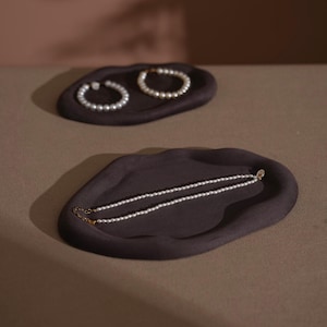 May include: Two dark brown, organic-shaped trays. One tray holds a pearl necklace, and the other holds two pearl bracelets. The jewelry is white and the trays are a dark brown color. The background is a neutral brown.