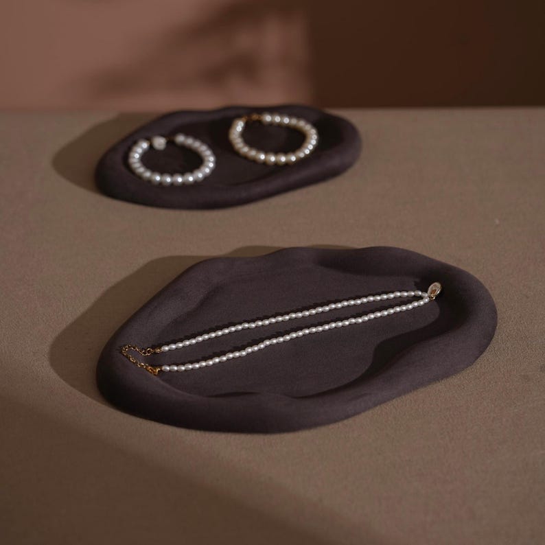 May include: Two dark brown, organic-shaped trays. One tray holds a pearl necklace, and the other holds two pearl bracelets. The jewelry is white and the trays are a dark brown color. The background is a neutral brown.