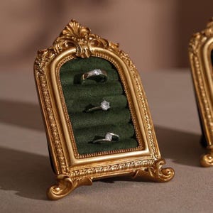 May include: A gold-colored, ornate picture frame-style ring holder with a dark green velvet interior. Three rings with clear stones are displayed. The frame has a decorative, vintage design.