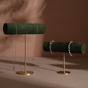 May include: Two jewellery display stands. The stands have a dark green, velvet-covered horizontal bar supported by a gold-coloured metal stem and base. One stand displays two gold necklaces, and the other displays two bracelets.