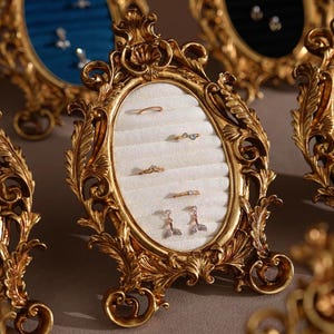 May include: Ornate gold-colored jewelry display frame with an oval center. The frame holds several pieces of jewelry, including rings and earrings, against a white, velvety background. The frame has a detailed, vintage design.