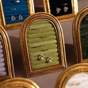 May include: Gold-framed jewelry display with a green velvet interior. The display holds rings and earrings. The frame has an arched top and a decorative border. Other displays with blue and green velvet interiors are visible.