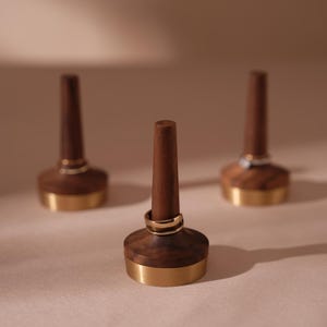 May include: Three wooden ring holders with a dark brown finish and gold-coloured metal bases. One holder displays a gold ring. The holders have a cylindrical post for displaying rings. The background is a soft, neutral colour.