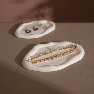 May include: Two off-white, organically shaped jewelry trays. One holds gold earrings, the other a gold necklace with pearls and small crystals. The trays sit on a neutral-toned surface, with a soft, diffused light.